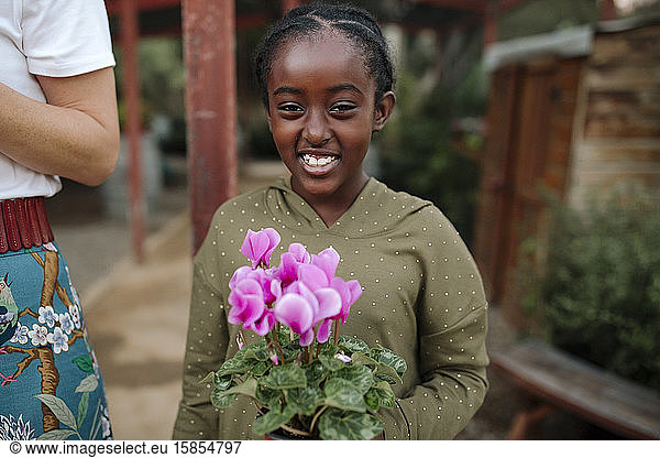 Smiling black girl holding plant with pink flowers at nursery