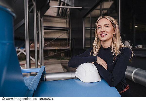 Smiling blond businesswoman leaning on metal structure in factory