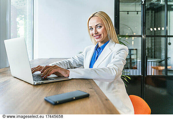 Smiling blond businesswoman with laptop sitting at desk in office