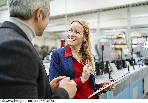 Smiling blond businesswoman with tablet PC looking at colleague discussing in factory