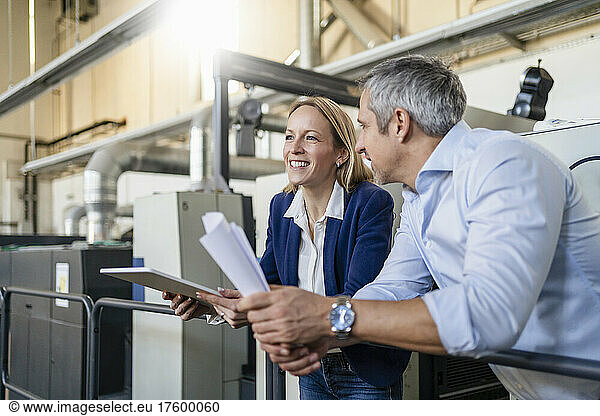 Smiling blond businesswoman with tablet PC standing by colleague in factory