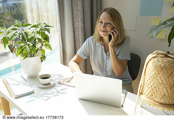 Smiling blond freelancer talking on smart phone sitting at desk in home office
