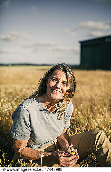 Smiling blond woman holding flowers while sitting at agricultural filed during sunny day