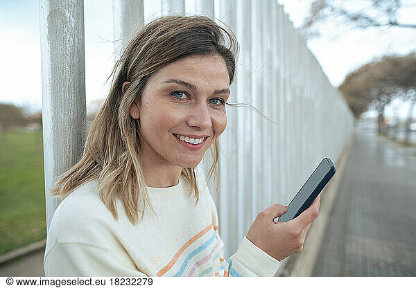 Smiling blond woman holding smart phone leaning on fence