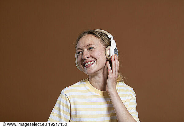 Smiling blond woman listening music through wireless headphones over brown background