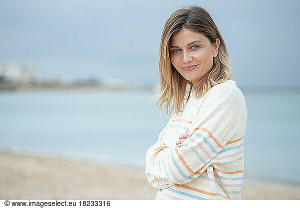 Smiling blond young woman with arms crossed standing at beach