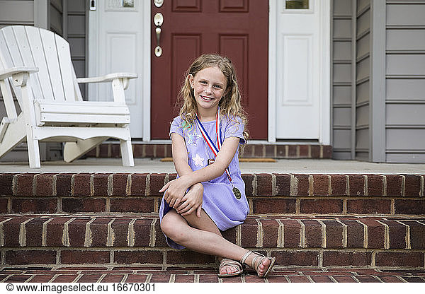 Smiling Blonde Girl Wearing A Medal Sits on Front Brick Front Steps
