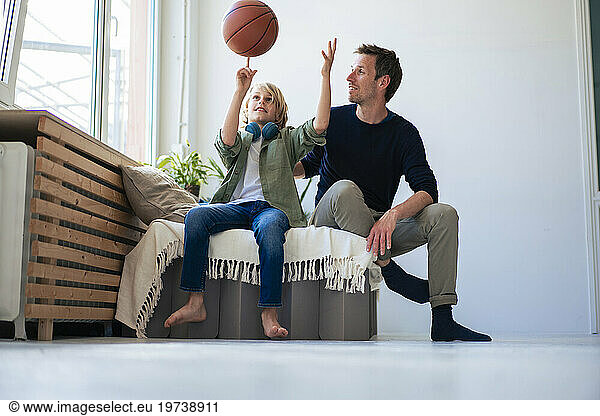 Smiling boy balancing basketball on finger with father sitting at home