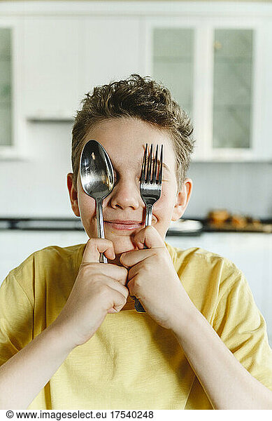 Smiling boy covering eyes with fork and spoon at home