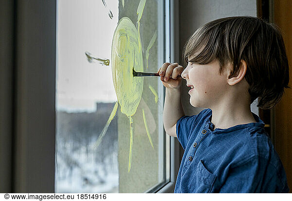Smiling boy drawing sun on glass with paint brush