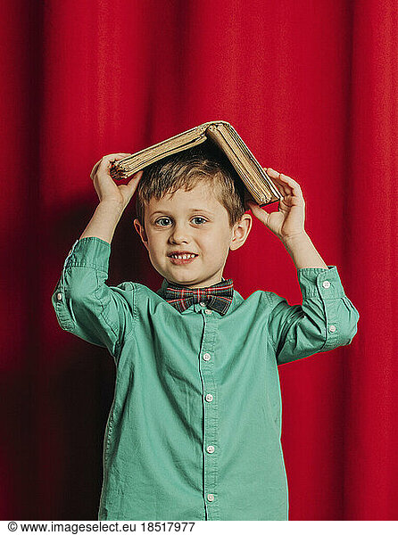 Smiling boy holding book on head in front of red curtain