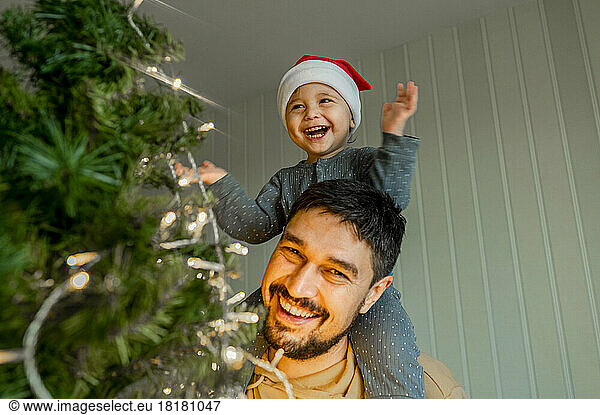 Smiling boy sitting on father's shoulder having fun decorating Christmas tree at home