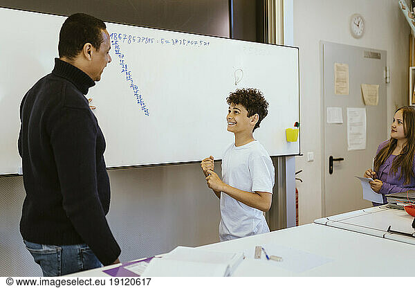 Smiling boy talking to male teacher standing near whiteboard in classroom