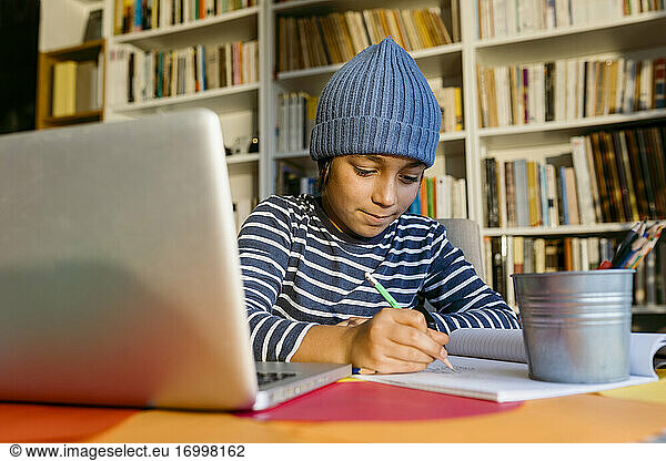 Smiling boy wearing knit hat writing in book while sitting at home