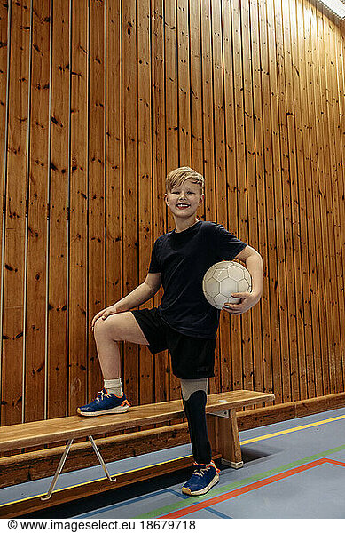 Smiling boy with amputated leg holding soccer ball while standing by wooden wall at sports court