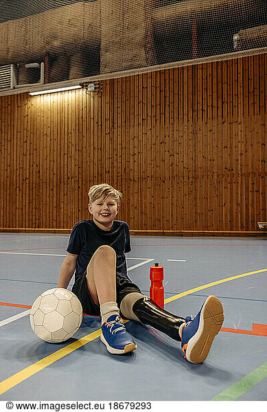 Smiling boy with amputated leg sitting on floor at football sports court