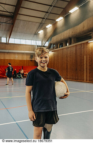 Smiling boy with disability holding soccer ball while standing at sports court