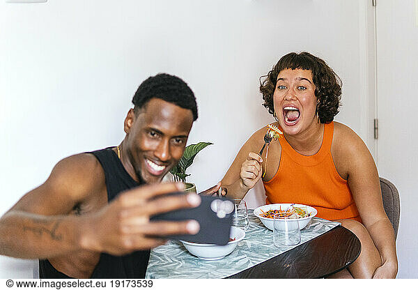 Smiling boyfriend taking selfie with girlfriend at dining table in kitchen