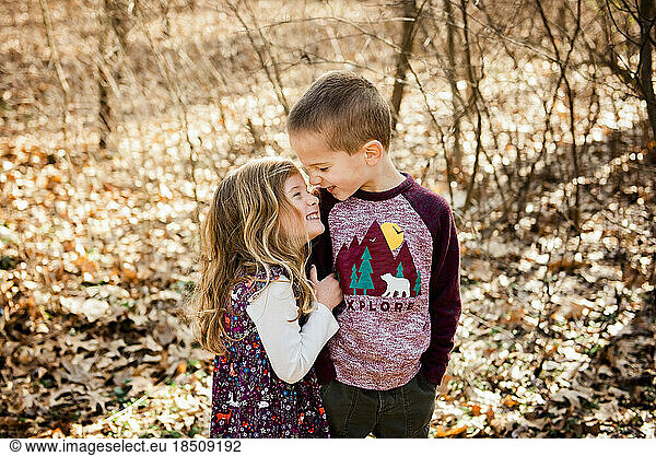 Smiling brother and sister stand in fall leaves with noses together