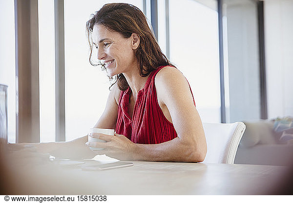 Smiling brunette woman drinking coffee and working at dining table