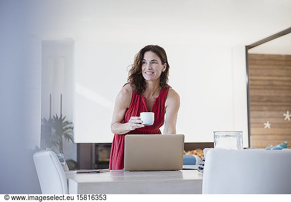 Smiling brunette woman drinking coffee  working at laptop at dining room table