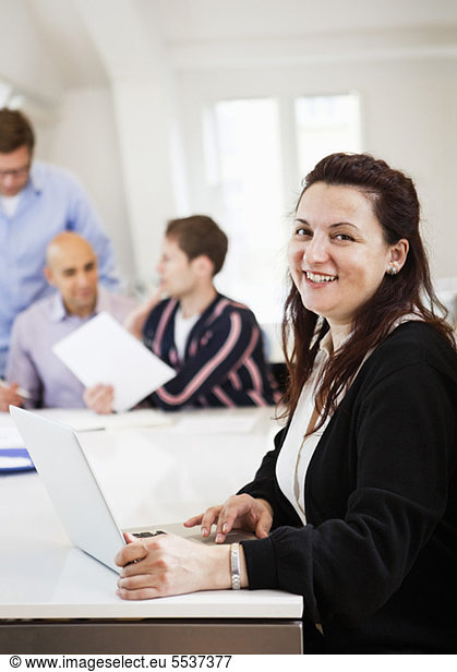Smiling business woman using laptop with colleagues at conference table in background