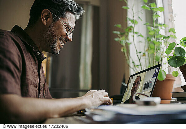 Smiling businessman discussing with male entrepreneur while working on laptop at home during COVID-19