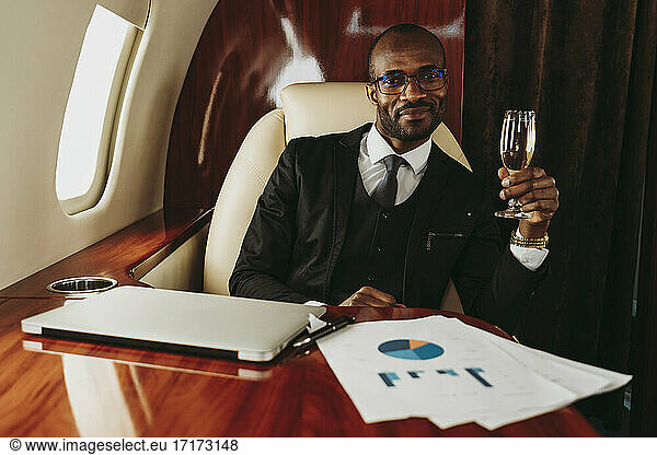 Smiling businessman holding champagne by laptop and document in airplane