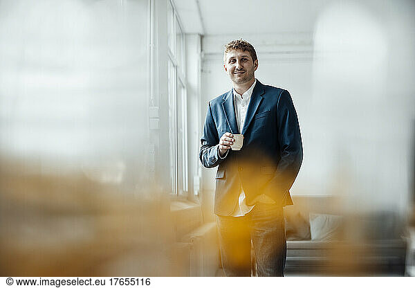 Smiling businessman holding coffee cup standing with hand in pocket at office
