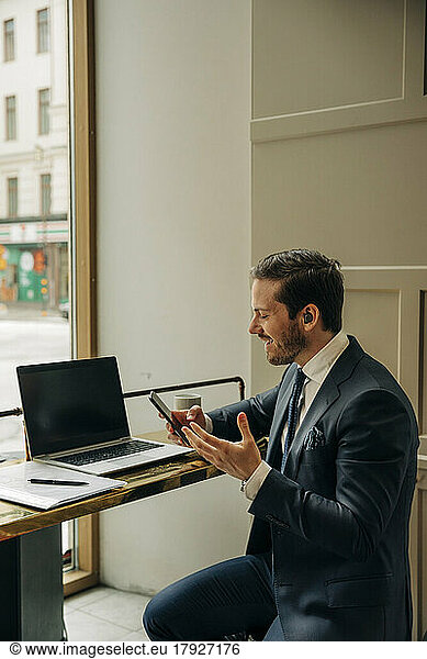 Smiling businessman holding mobile phone while talking through wireless-in ear headphones at office