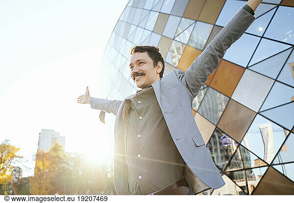 Smiling businessman in suit with arms outstretched by modern office building on sunny day