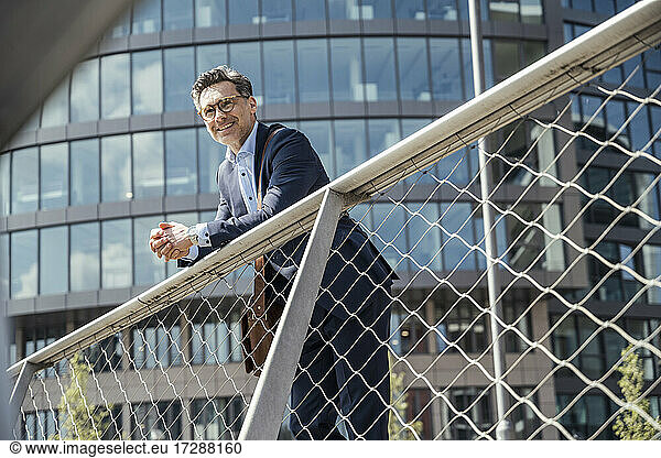 Smiling businessman leaning on railing during sunny day