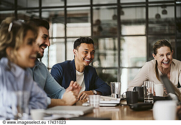 Smiling businessman looking away while sitting amidst cheerful colleagues at conference table