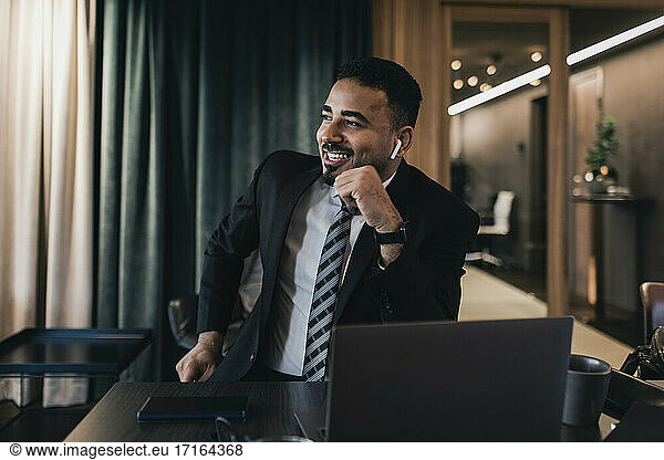 Smiling businessman looking away while talking through wireless in-ear headphones in board room