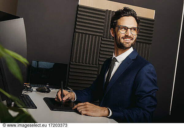 Smiling businessman looking away while using graphics tablet at desk in office