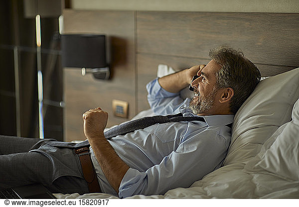 Smiling businessman lying on the bed in hotel room talking on the phone