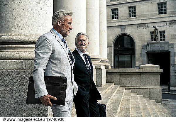 Smiling businessman moving down on steps with colleague near columns