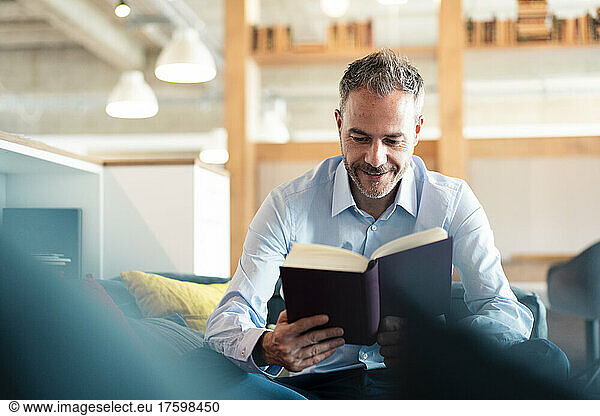 Smiling businessman reading book sitting at workplace