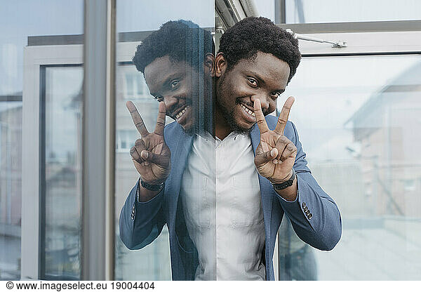 Smiling businessman showing peace sign peeking from glass window at office
