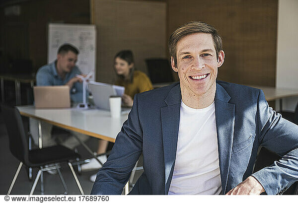 Smiling businessman sitting on chair in office with colleagues in background
