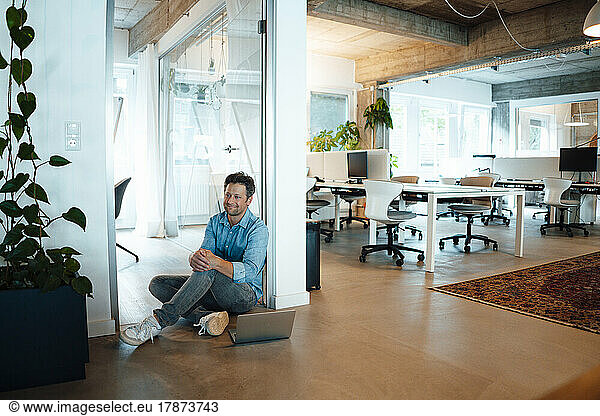 Smiling businessman sitting on floor by laptop in office