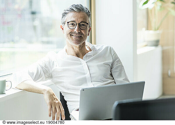 Smiling businessman sitting with laptop leaning head on window sill at office