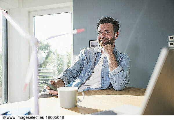 Smiling businessman sitting with mobile phone and hand on chin at desk
