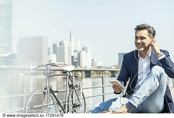 Smiling businessman sitting with mobile phone and in-ear headphones during sunny day