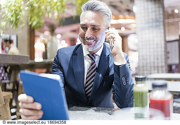 Smiling businessman talking on smart phone holding tablet PC at cafe