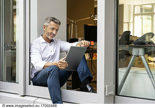 Smiling businessman using digital tablet while sitting at office door