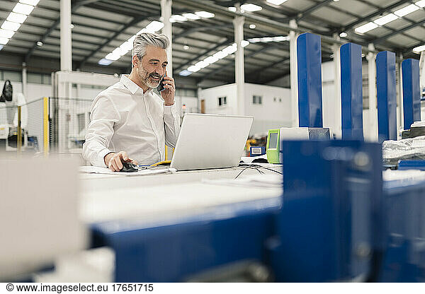 Smiling businessman using laptop talking on mobile phone at desk in factory
