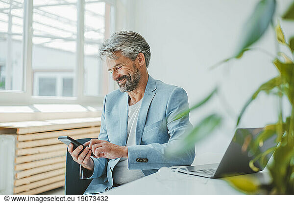 Smiling businessman using mobile phone sitting at desk in office