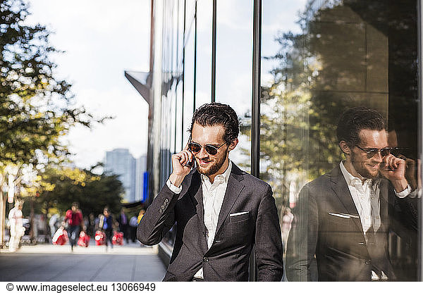 Smiling businessman using phone reflecting on glass building in city