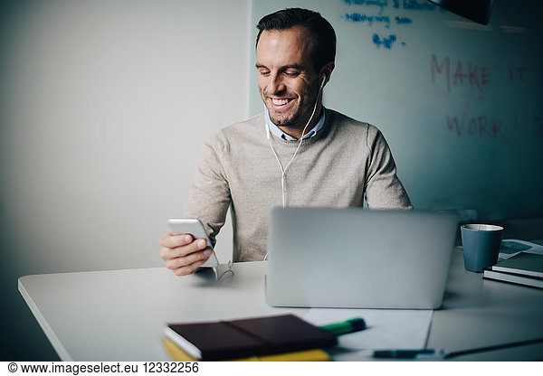 Smiling businessman using smart phone and laptop while sitting at desk in creative office